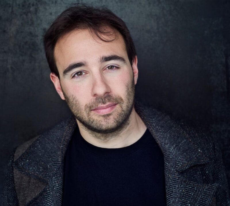 Man with short hair and a beard, wearing a dark coat, stands against a dark textured backdrop.