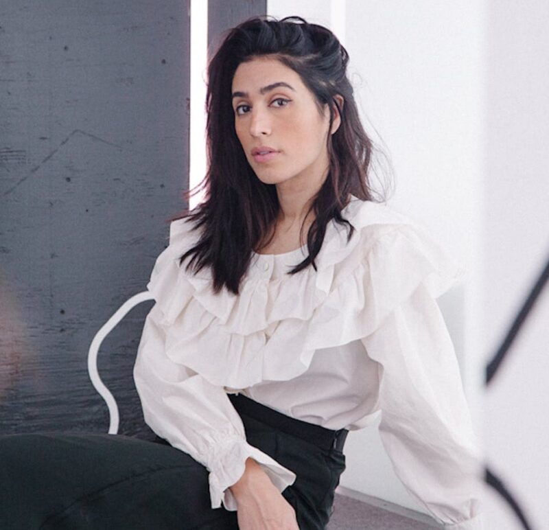 Woman in a white ruffled blouse and black skirt sits in a modern setting with abstract black and white designs in the background.