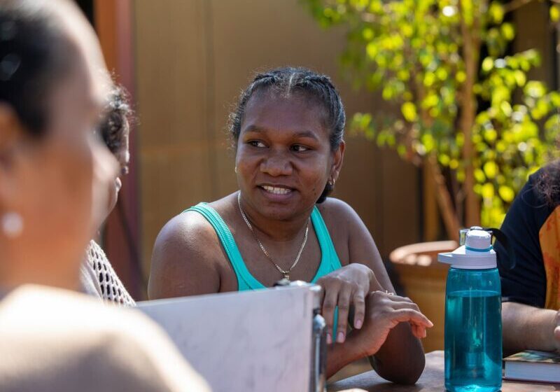 A Black woman in a teal shirt, sits around a table of women having a conversation under the sun.