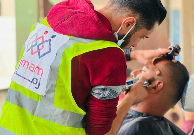 A young Palestinian man with a yellow vest adorning the Zimam logo, shaves another mans hair.