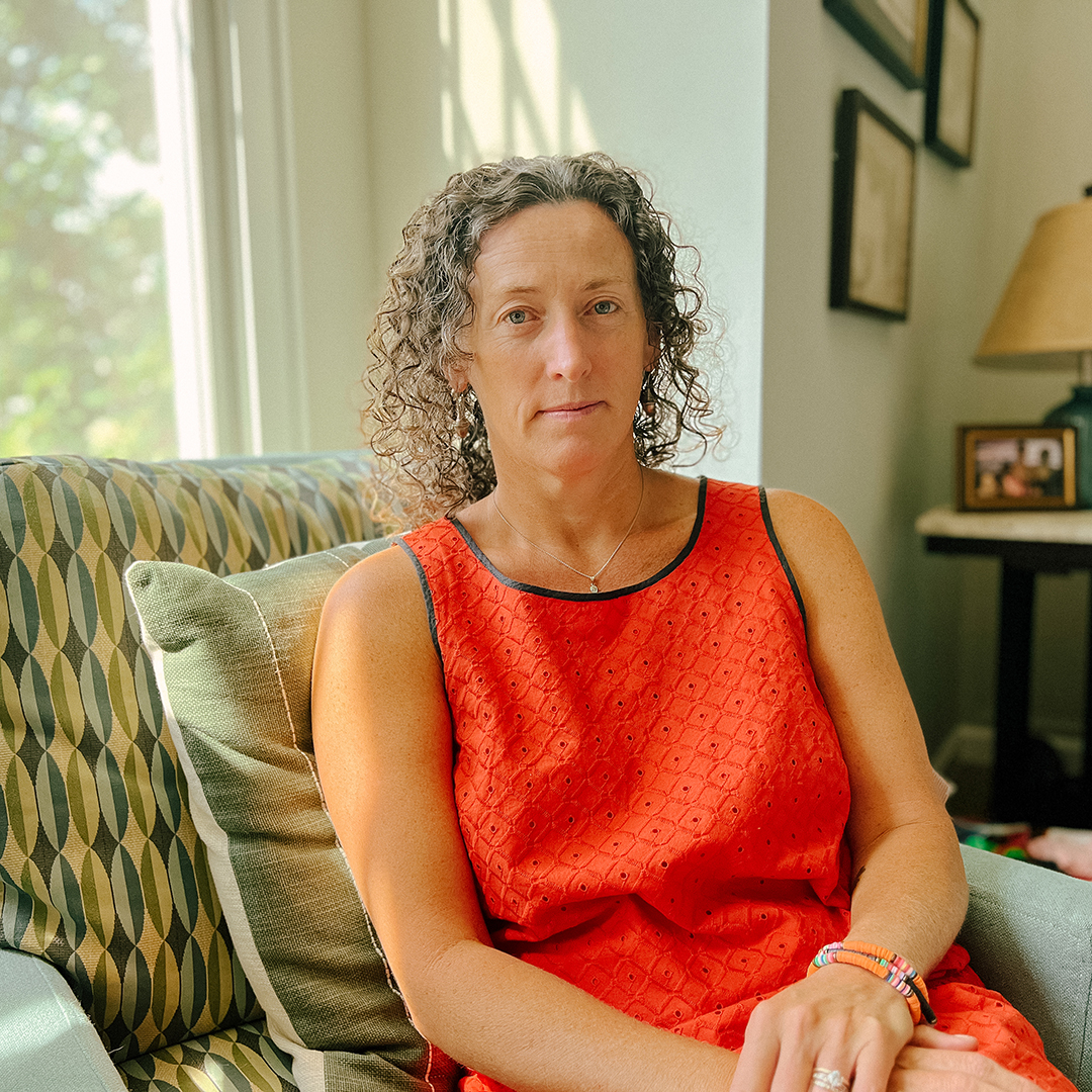 A person with curly hair in a red dress sits on a patterned chair near a window, with sunlight streaming in.