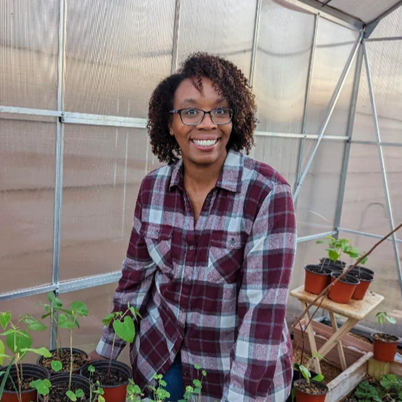 A person in a plaid shirt smiles while standing in a greenhouse surrounded by potted plants.