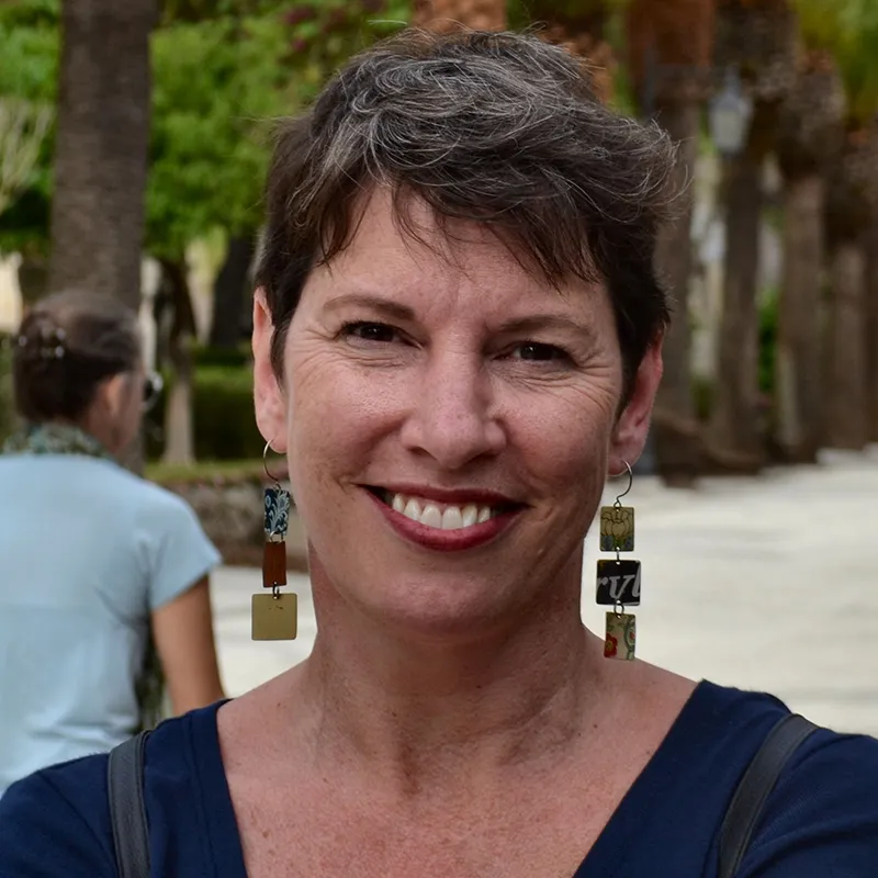 Smiling woman with short hair, wearing colorful rectangular earrings, stands outside with trees in the background.