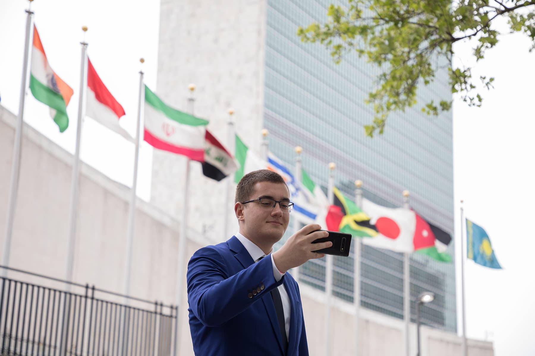 A young man in glasses and a blue suit takes a selfie in front of international flags.
