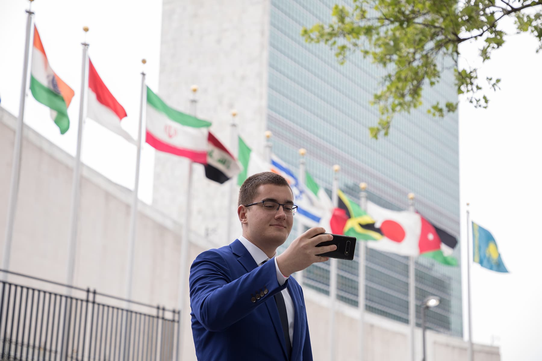 A young man in glasses and a blue suit takes a selfie in front of a building adorned with international flags.