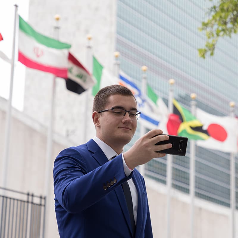 A young man in glasses and a blue suit takes a selfie in front of international flags.