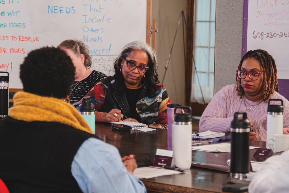 A group of diverse women sit around a table listening to one another in a group conversation.