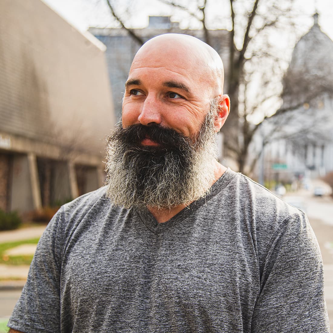 A man with a black and grey beard, standing outside in the sun in front of a legislative building.