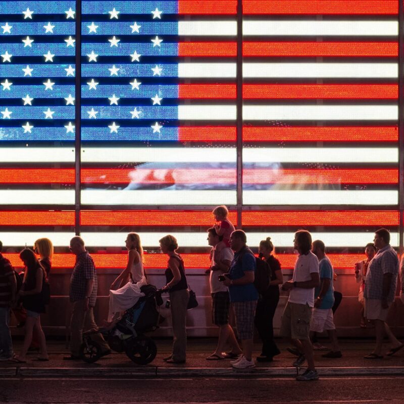 People walk in front of a large electronic display of the American flag at night.