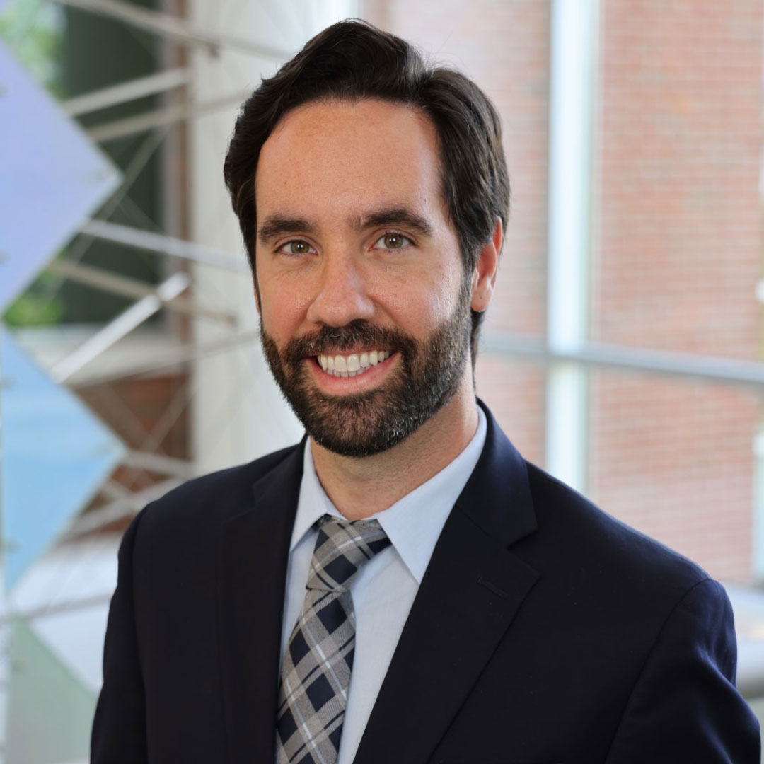 Smiling man with a beard and dark hair in a suit and checkered tie, standing indoors.