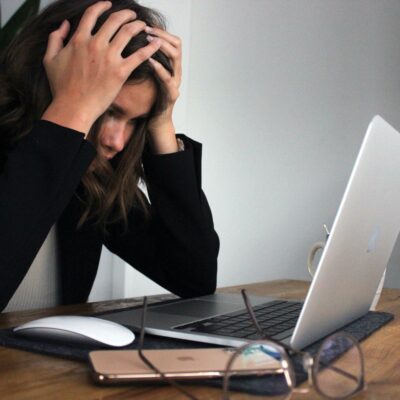 A person sits at a desk, holding their head in frustration while looking at a laptop. Glasses and a smartphone lie on the table.