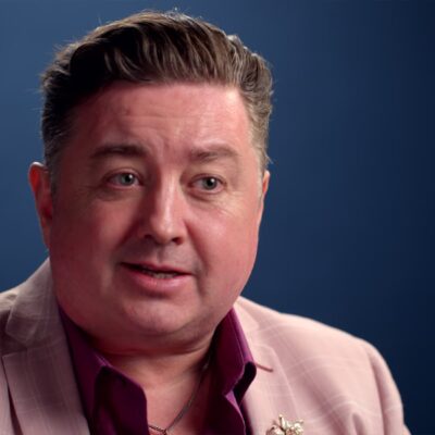 A person in a pink suit and maroon shirt speaks against a blue background.