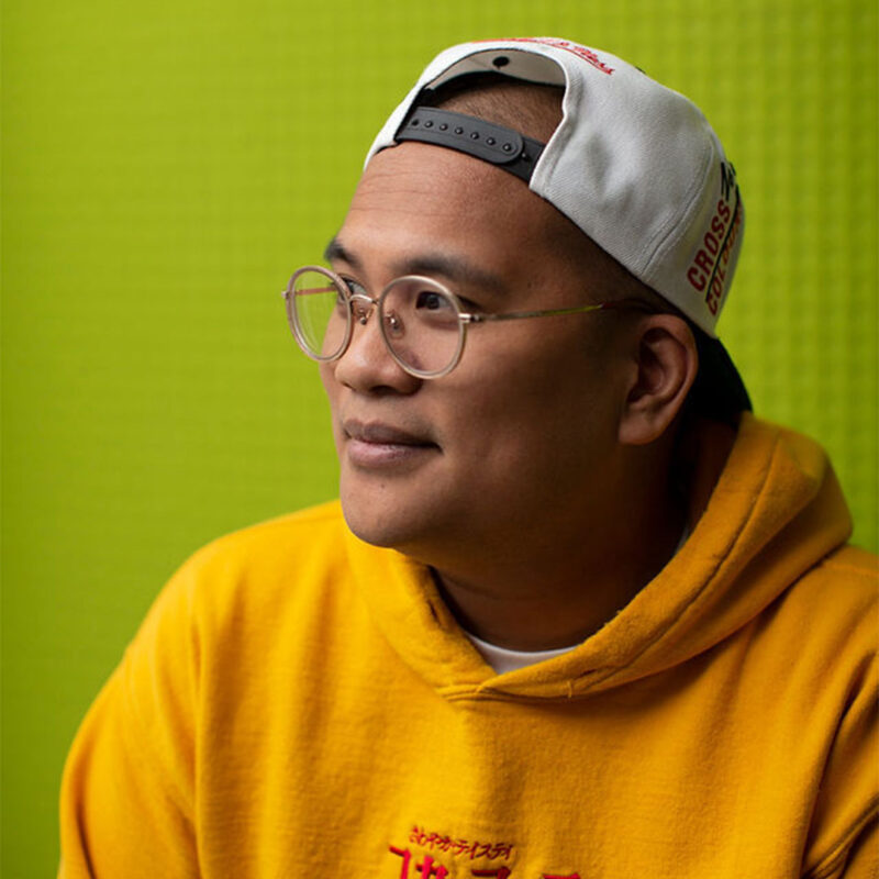 Person wearing glasses, a yellow hoodie, and a backward white cap, smiling against a bright green background.