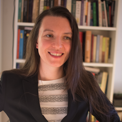 Smiling person with long hair wearing a blazer and striped top, standing in front of a bookshelf.