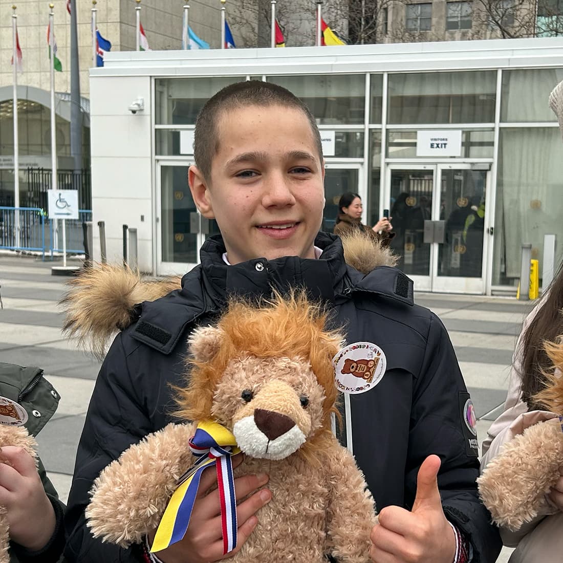 A child holds teddy bears with ribbons in front of a building with international flags.