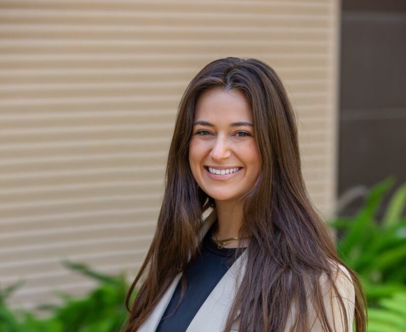 Woman with long brown hair smiling, wearing a beige blazer and black top, standing in front of a beige wall with greenery in the background.