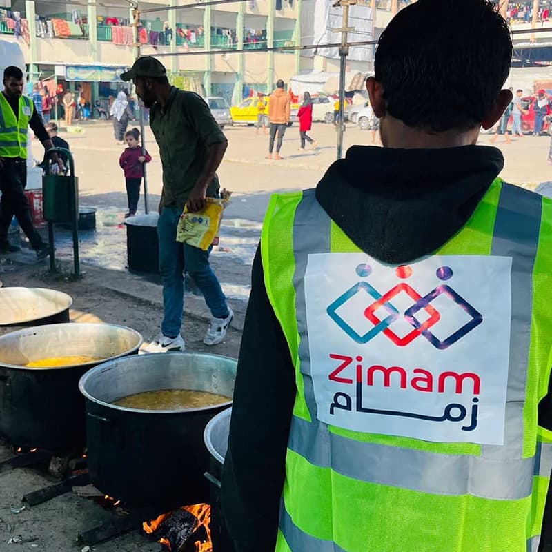 A young man standing in front of large pots of food. He is wearing a yellow vest that has the logo for Zimam on it.
