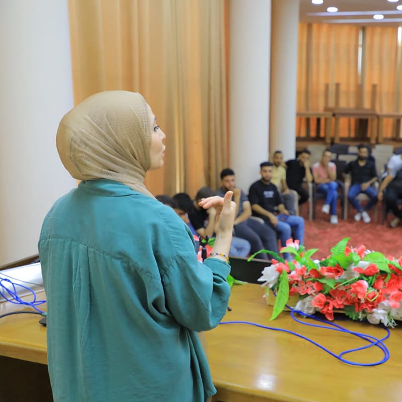 An Arabic woman wearing a teal shirt and beige headscarf, talking to a group of young people arranged in a circle.
