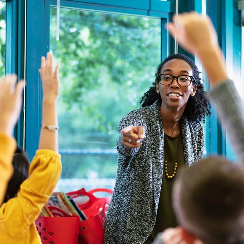 A teacher in a classroom pointing at a diverse group of children who have their hands raised.