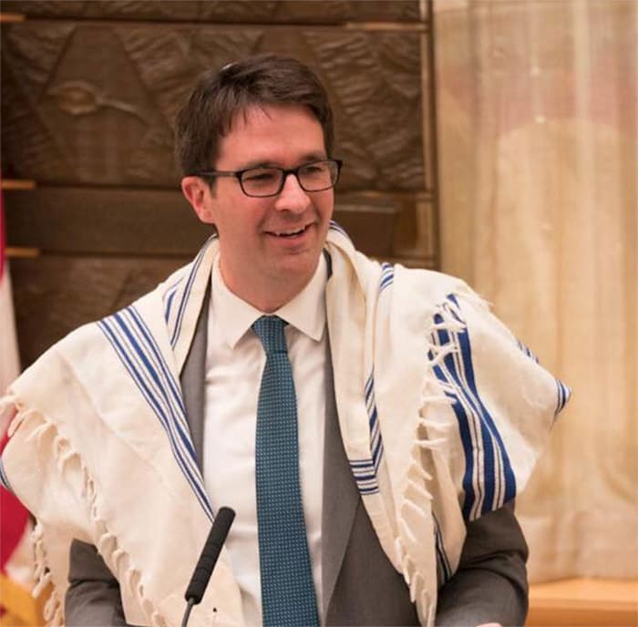 A man in a suit, wearing a striped shawl, is smiling and speaking at a podium.