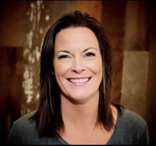 A smiling woman with dark hair wearing a gray top against a brown, textured background.