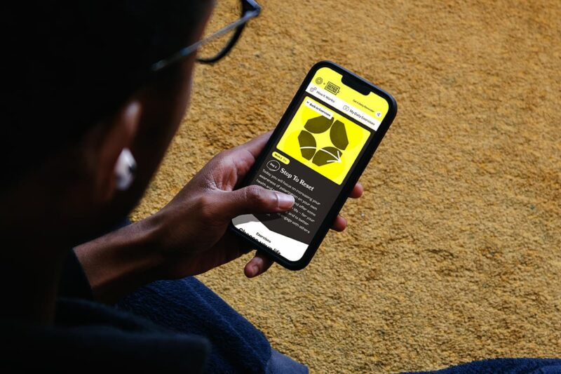 Person holding a smartphone displaying a yellow and black website with leaf graphics and text, sitting on a yellow carpet.