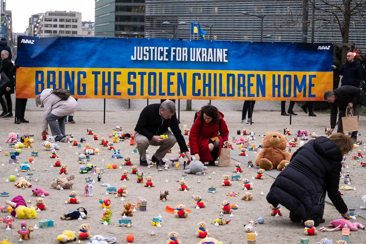 People placing small teddy bears on the ground in front of a sign that reads "Justice for Ukraine, Bring The Stolen Children Home"