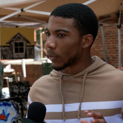 A person in a tan and white hoodie speaks into a microphone at an outdoor event with tents and artwork in the background.