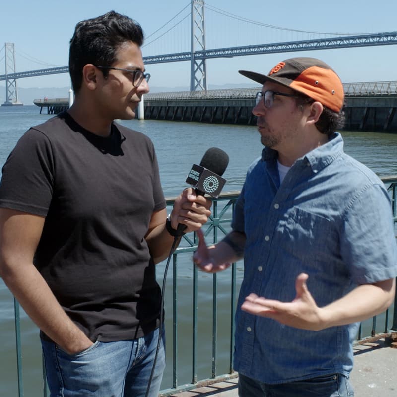 A man in a black t-shirt, interviewing a man in a denim shirt on camera, holding a microphone to him that says Starts With Us on it.